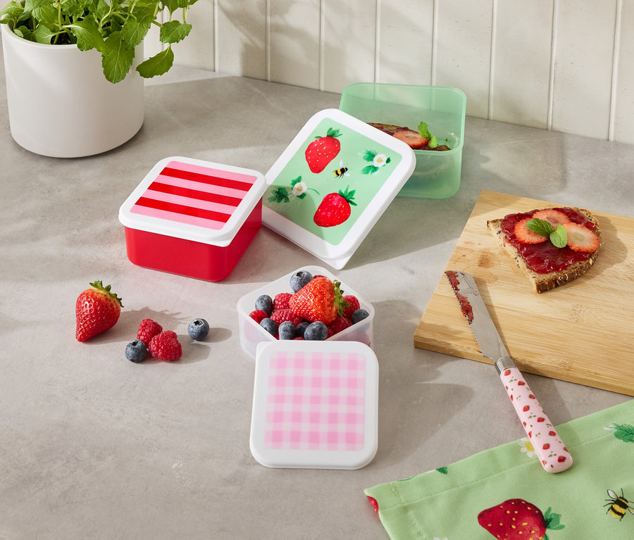 Na kuchynskej linke stojí niekoľko farebných nádob na potraviny, niektoré naplnené ovocím, vedľa krajca chleba a noža.