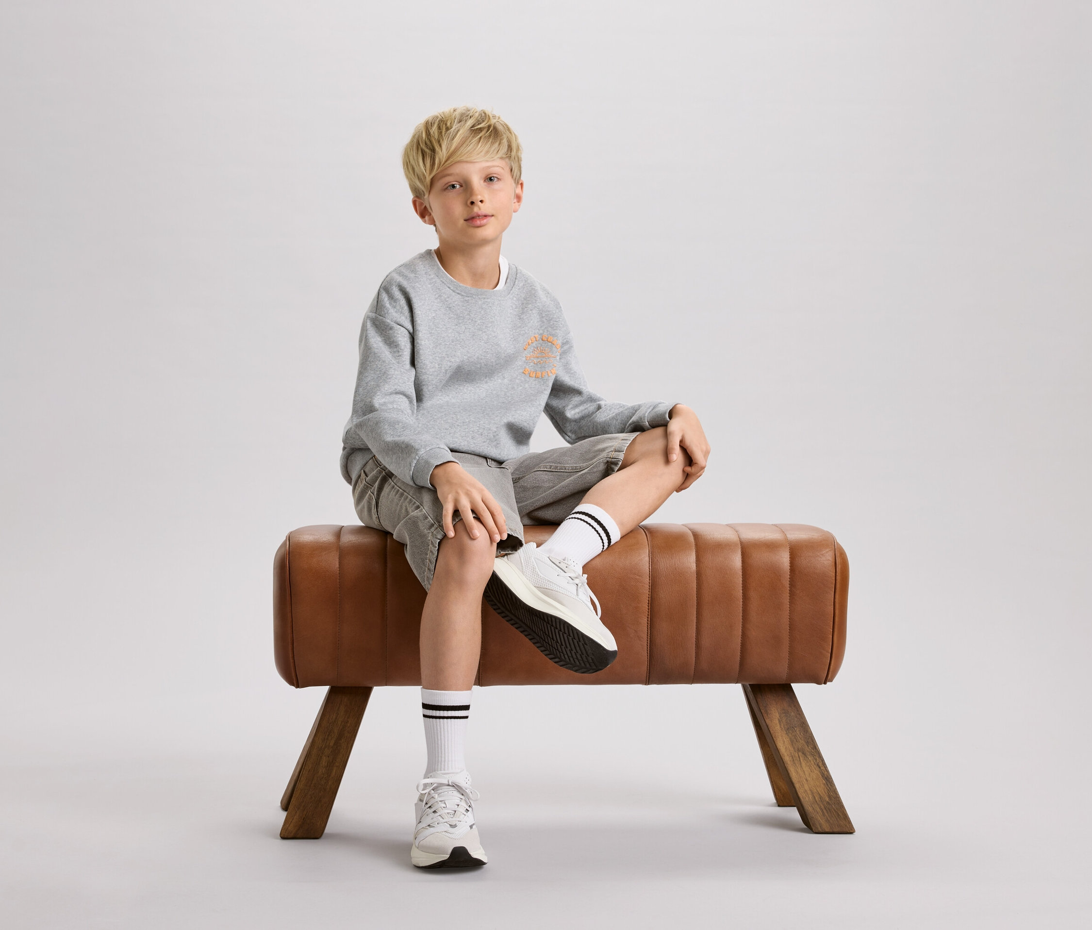 Chlapec s blond vlasmi sedí na hnedom gymnastickom koni. Má na sebe sivú mikinu, šortky a biele ponožky s čiernymi pruhmi.