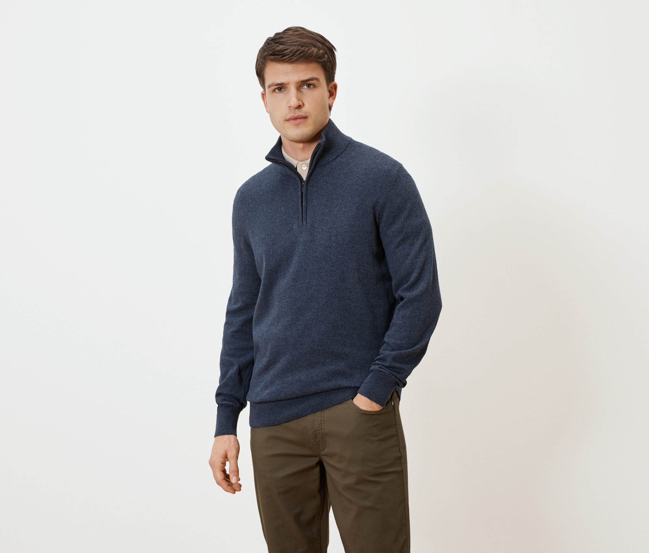 Muž v modrom svetri na zips a hnedých nohaviciach s rukou vo vrecku na neutrálnom pozadí.