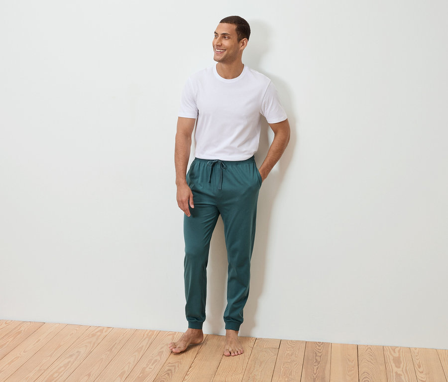 Muž stojí bosý s rukou vo vrecku pred bielou stenou. Má na sebe biele tričko a petrolejové tepláky.