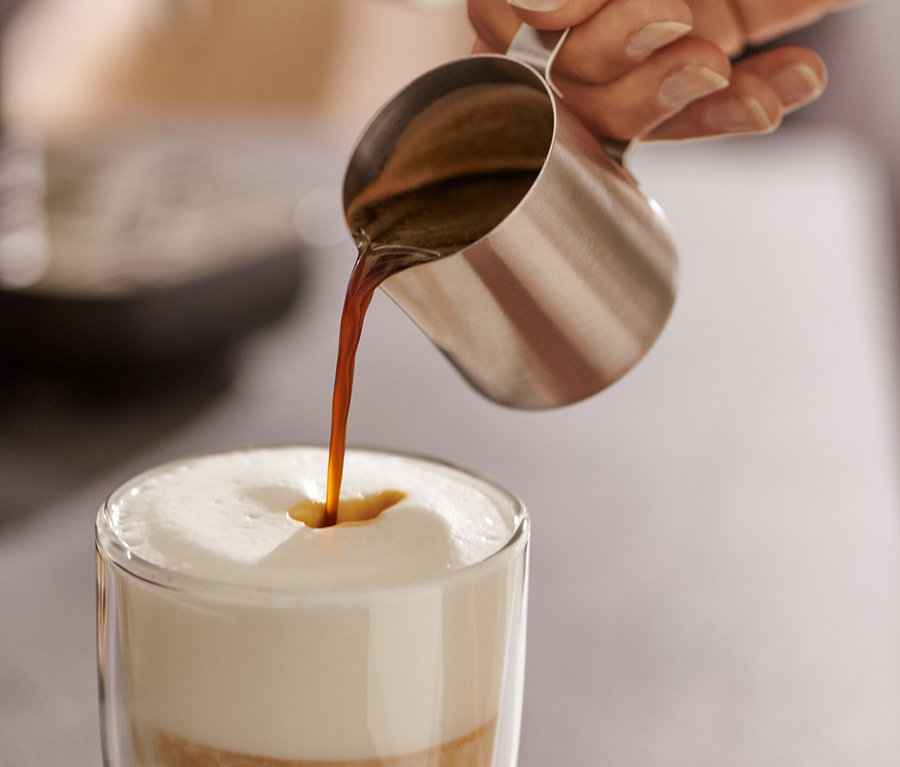Žena nalieva espresso z kanvičky z ušľachtilej ocele (80 ml) do pohára s mliekom.