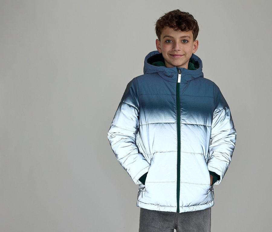 Usmievavý chlapec s kučeravými vlasmi má na sebe prešívanú bundu s kapucňou. Bunda má prechod farieb od tmavomodrej hore po reflexnú striebornú dole a zelený zips. Ruky má vo vreckách bundy.