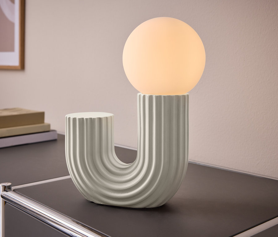 Stolová lampa v tvare U s rebrovanou základňou a guľovou svietiacou žiarovkou, umiestnená na tmavom stole. V pozadí sú knihy a obraz na stene.