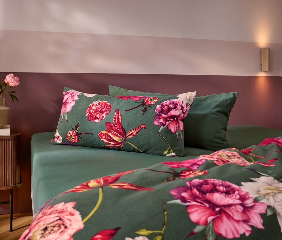 Posteľ s tmavozelenými obliečkami s veľkými ružovými a červenými kvetmi a jednofarebným zeleným vankúšom. Vľavo stojí nočný stolík s vázou plnou ružových pivoniek. Stena je dvojfarebná, ružová a tmavočervená.