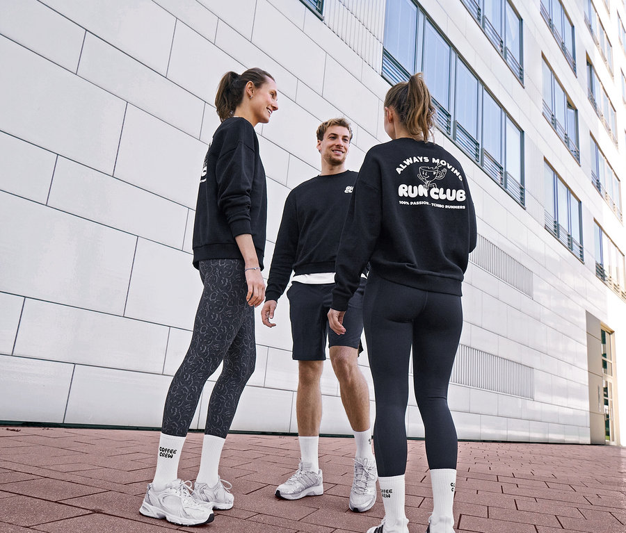 Traja ľudia v čiernych mikinách a športovom oblečení sa rozprávajú. Žena má na sebe sivo-čierne športové legínsy »ActivePower« a unisex ponožky Coffee Crew. Muž má na sebe čierne športové šortky a biele ponožky. Druhá žena má na sebe čierne športové legíny »ActivePower« a mikinu s logom „RUN CLUB“.