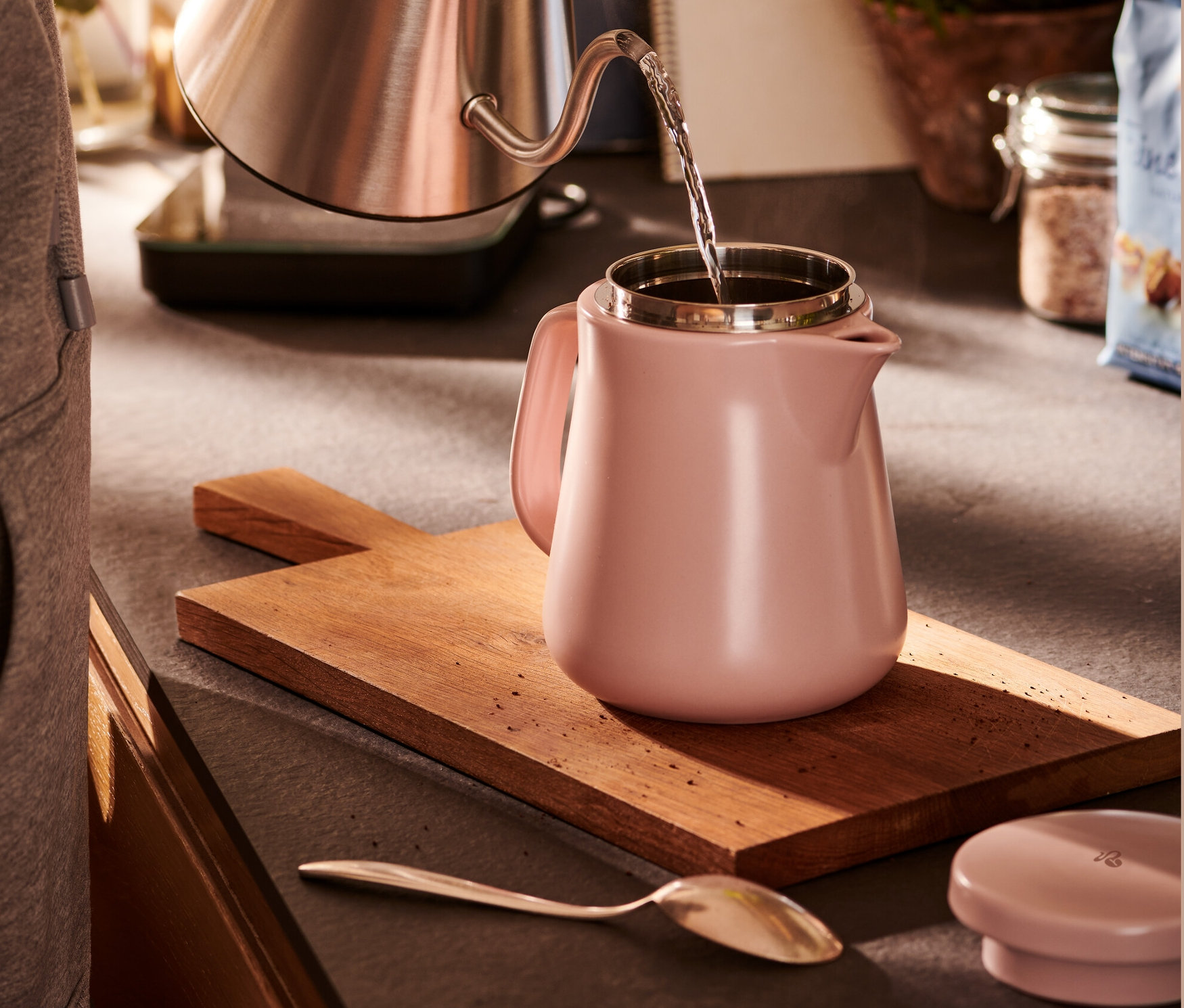 Žena nalieva vodu z nerezovej kanvice do keramickej kanvice s filtrom ružovej farby.