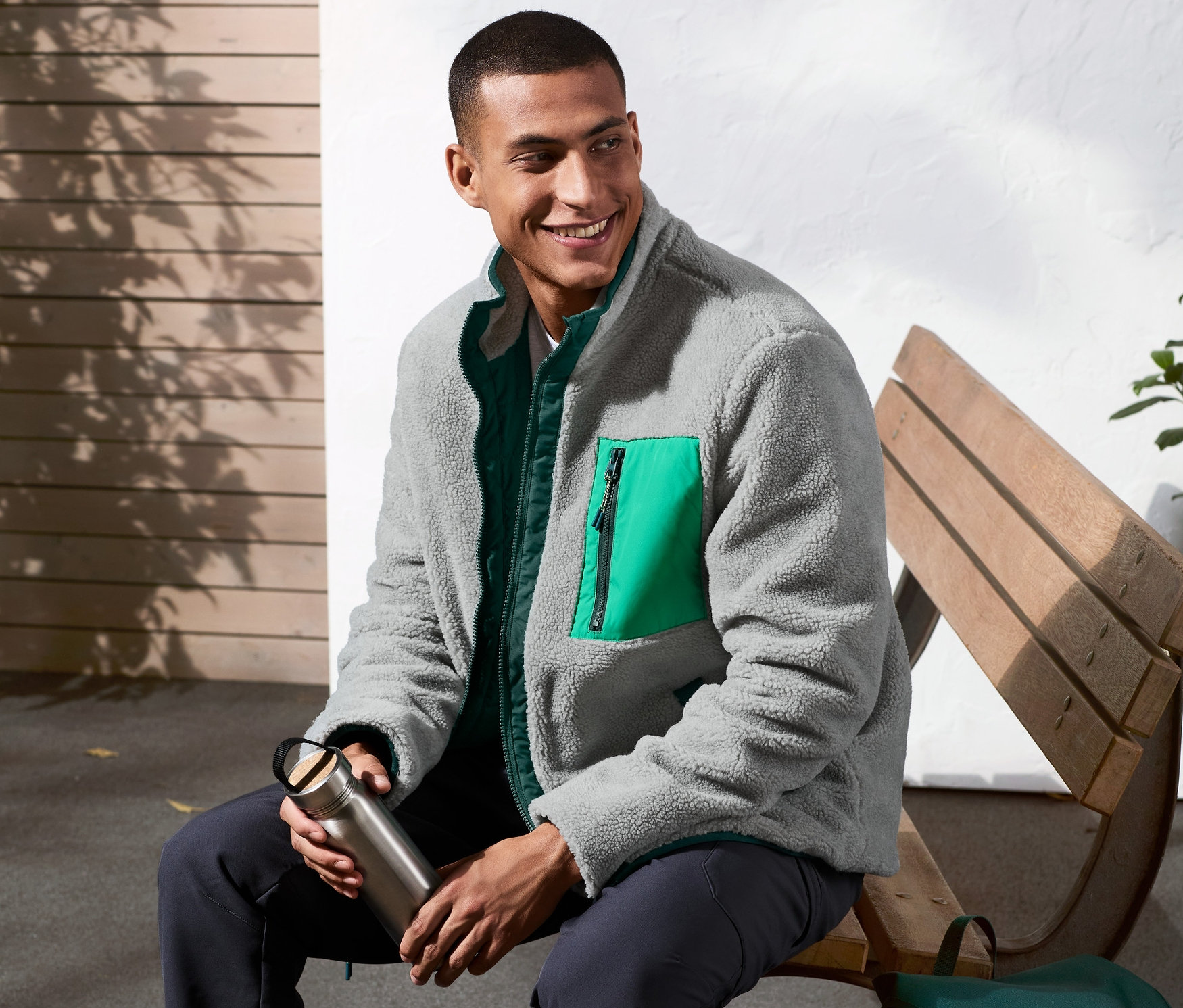 Muž sedí na lavičke v obojstrannej bundě, drží fľašu s vodou. Vedľa neho je zelený batoh.