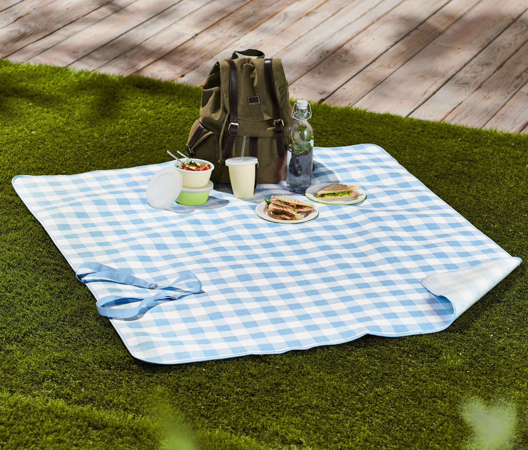 Na tráve leží malá kockovaná deka na piknik s jedlom, pitím a batohom.