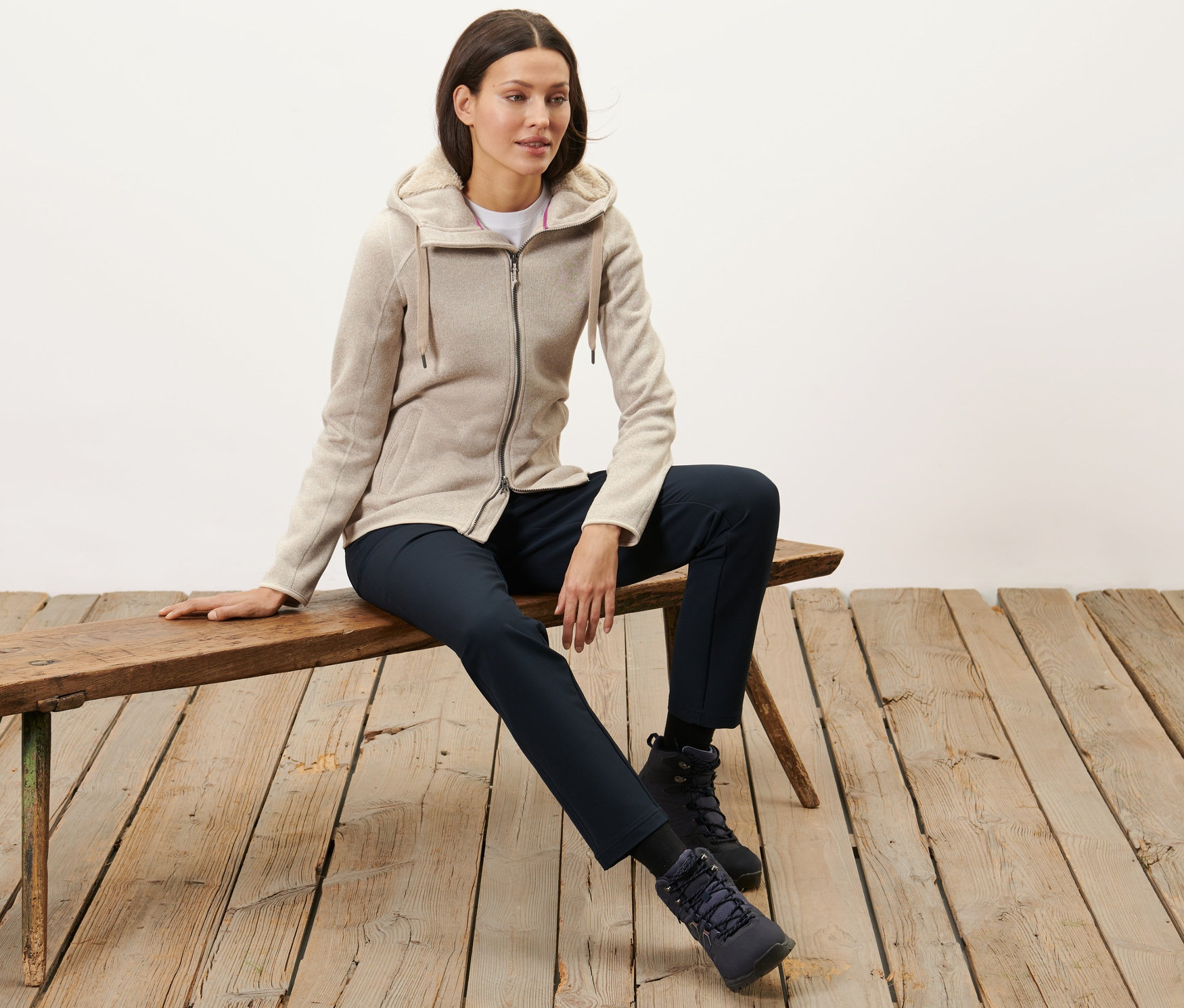 Žena sedí na lavičke a má na sebe pieskovú bundu z pleteného flísu s kapucňou.