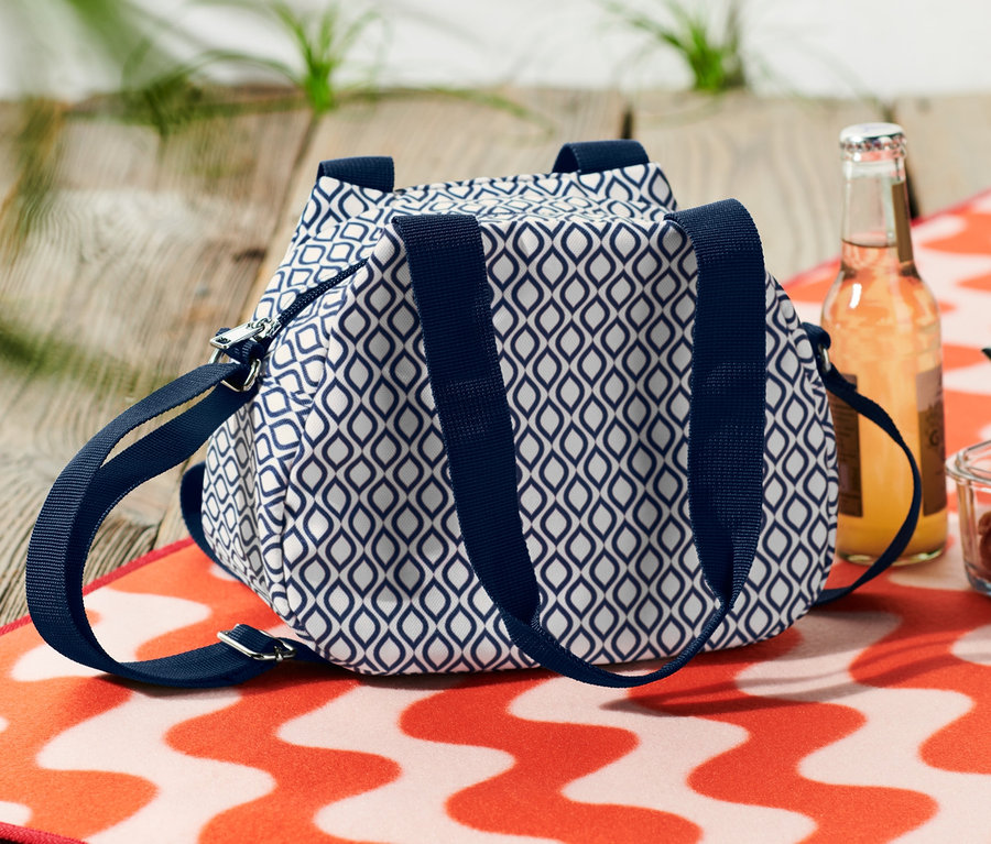 Modro-biela mini chladiaca taška leží vedľa nápoja a misky s cestovinami na oranžovo-bielej vlnitej plážovej osuške.