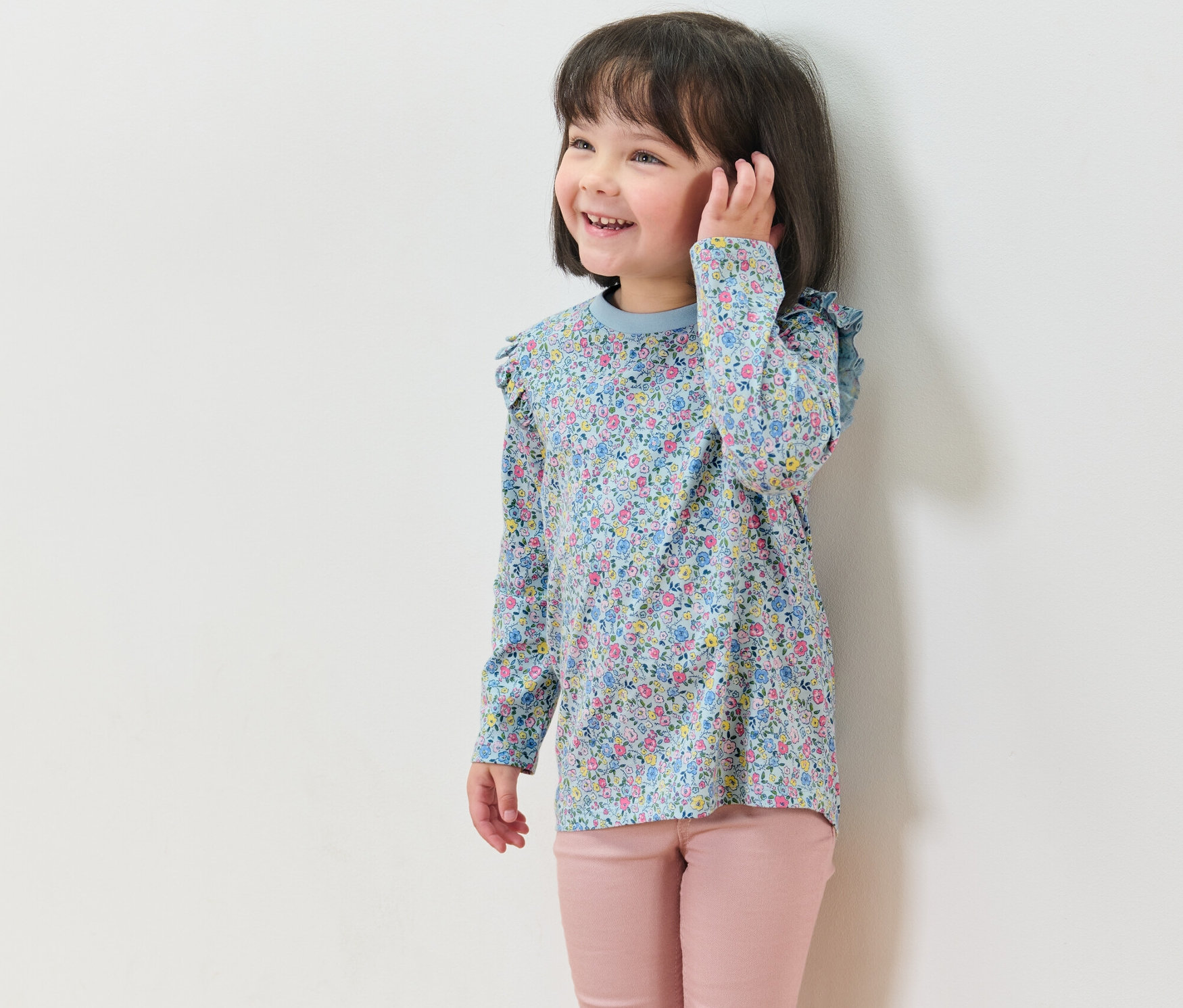 Usmievajúce sa dievča má na sebe kvetinové tričko s dlhým rukávom z 2-dielnej súpravy a ružové nohavice.