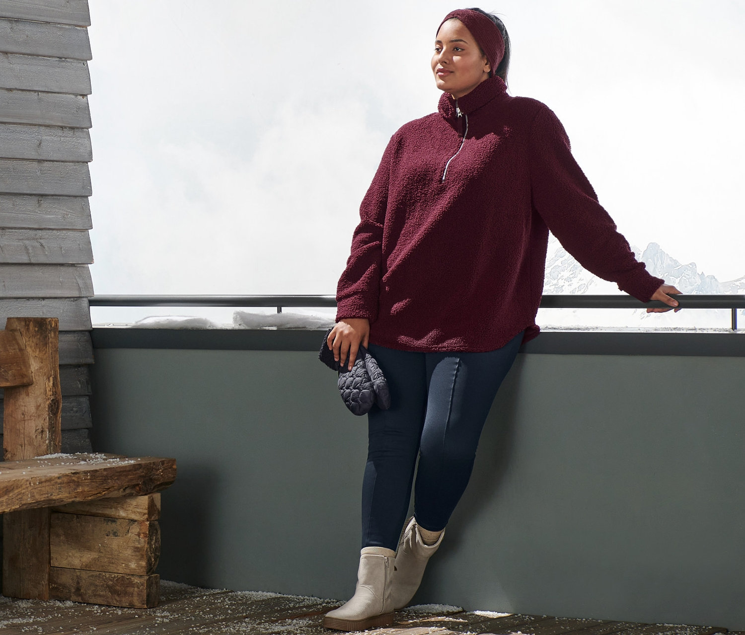 Žena opretá o zábradlie má na sebe plyšový pulóver v oversize štýle, bordovočervenú pletenú čelenku, modré termolegínsy a topánky z velúrovej kože. V ruke drží prešívané palčiaky.