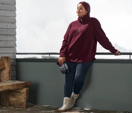 Žena opretá o zábradlie má na sebe plyšový pulóver v oversize štýle, bordovočervenú pletenú čelenku, modré termolegínsy a topánky z velúrovej kože. V ruke drží prešívané palčiaky.