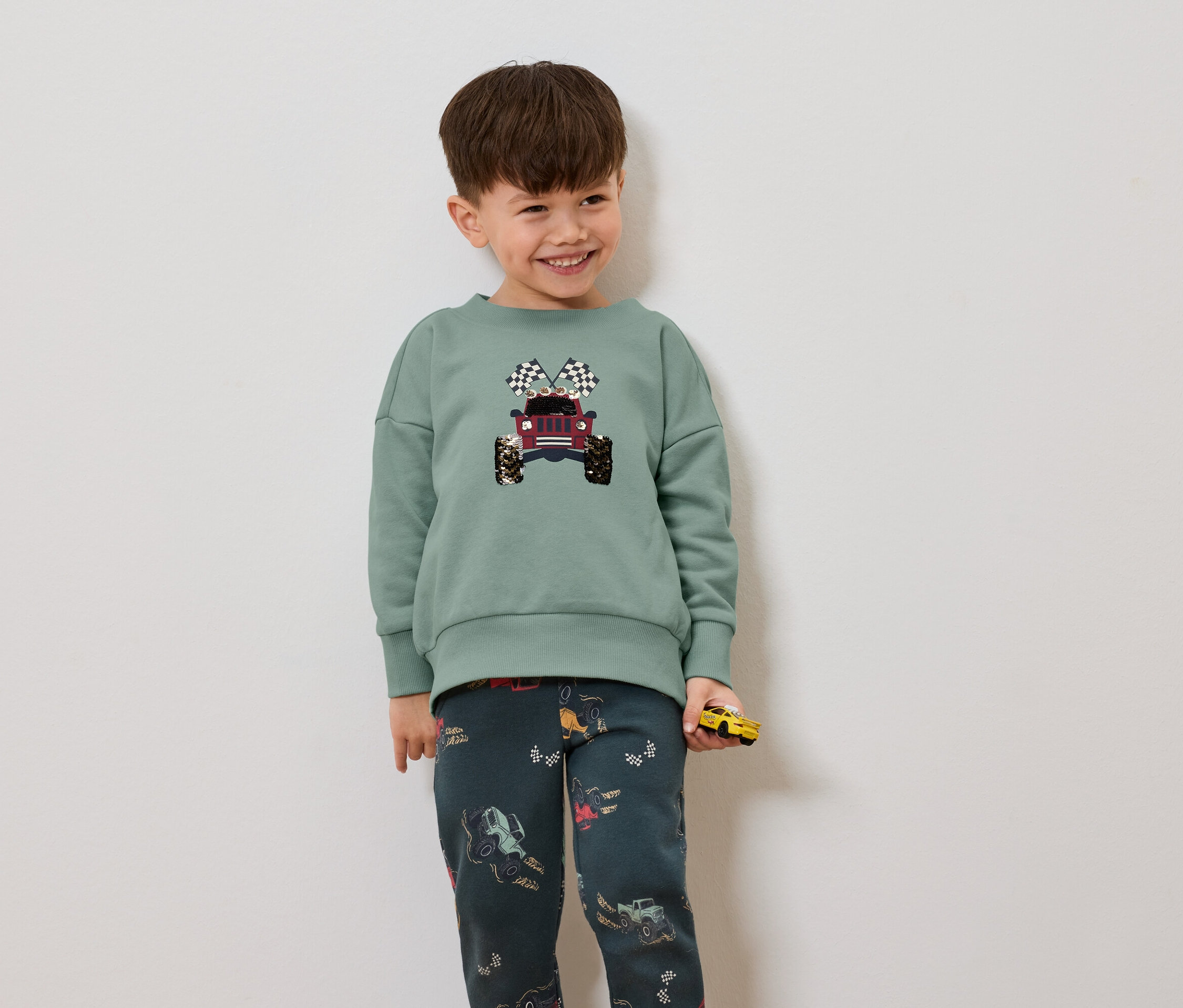 Chlapec v zelenom svetri s potlačou džípu a šachovnicovými vlajkami, má na sebe tmavé nohavice s potlačou džípov a drží žlté autíčko.