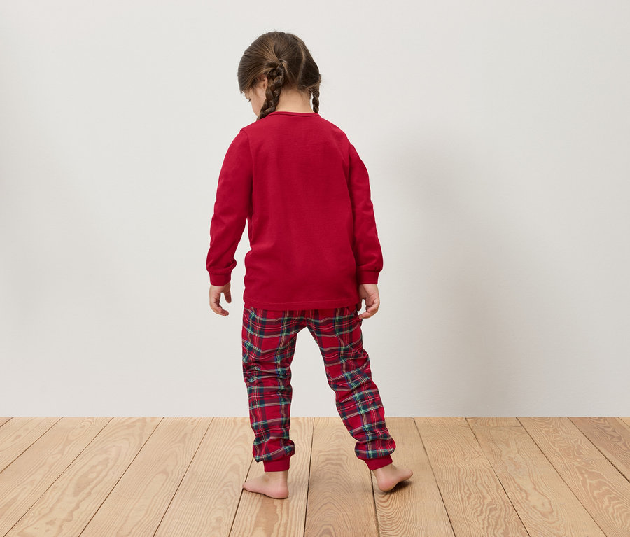 Dievča v červenom pyžame s dlhým rukávom a kockovaných nohaviciach stojí chrbtom k divákovi na drevenej podlahe.