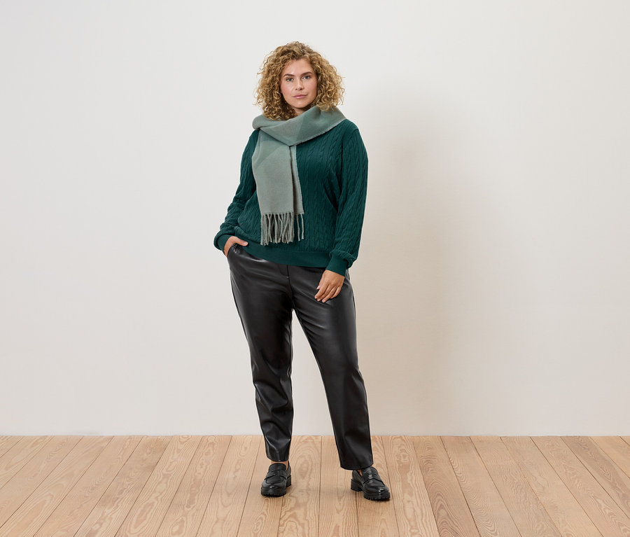 Žena s kučeravými vlasmi pózuje v zelenom svetri, šále a čiernych kožených nohaviciach.