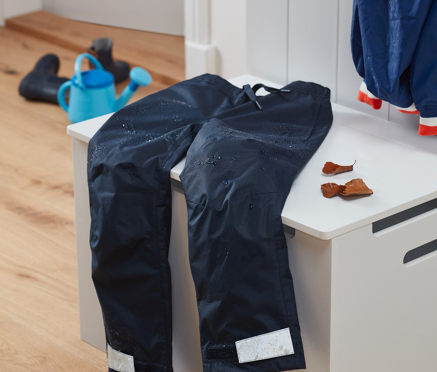 Tmavomodré nohavice do dažďa z recyklovaného materiálu ležia na bielej lavici. V pozadí je modrá kanva a gumáky.