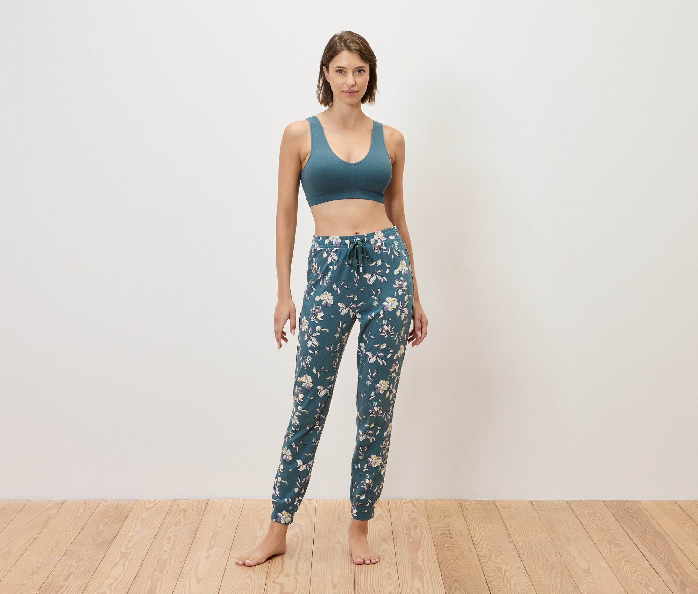 Žena stojí bosá pred stenou a má na sebe modrú športovú podprsenku a modré tepláky s kvetinovou potlačou.