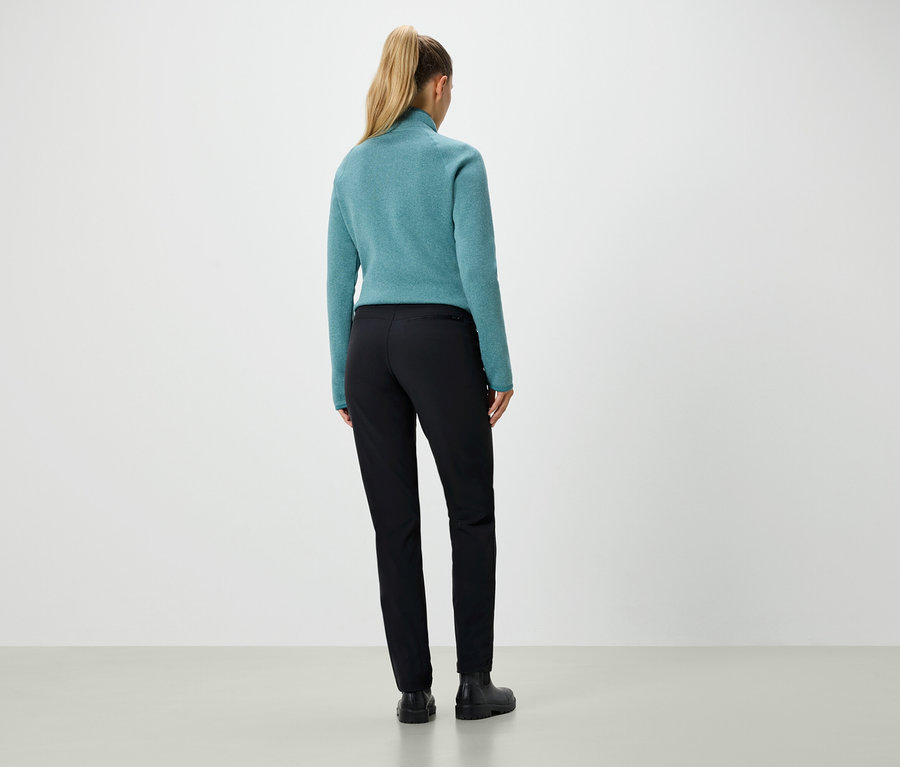 Pohľad zozadu na ženu s blond vlasmi zopnutými do copu, ktorá má na sebe modrozelený sveter a čierne nohavice.
