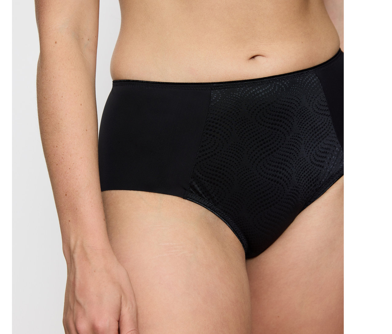 Osoba má na sebe čierne nohavičky Triumph Essential Minimizer Hipster X s hladkými bokmi a textúrovaným, vzorovaným predným dielom.