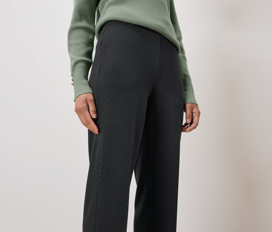 Žena má na sebe zelený sveter a čierne vzorované nohavice.