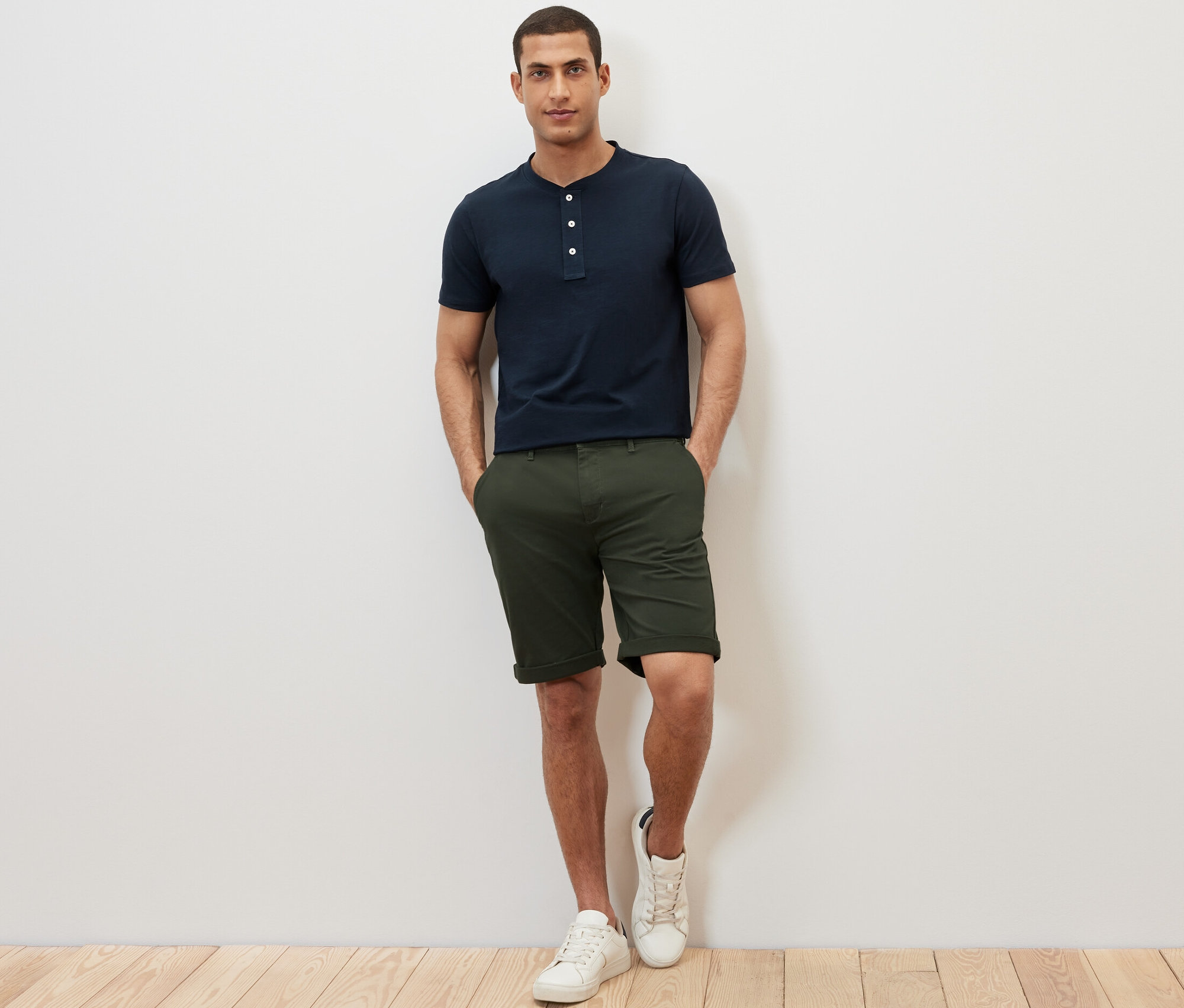 Muž stojí a má ruky vo vreckách šortiek. Má na sebe námornícke modré henley tričko s krátkymi rukávmi.
