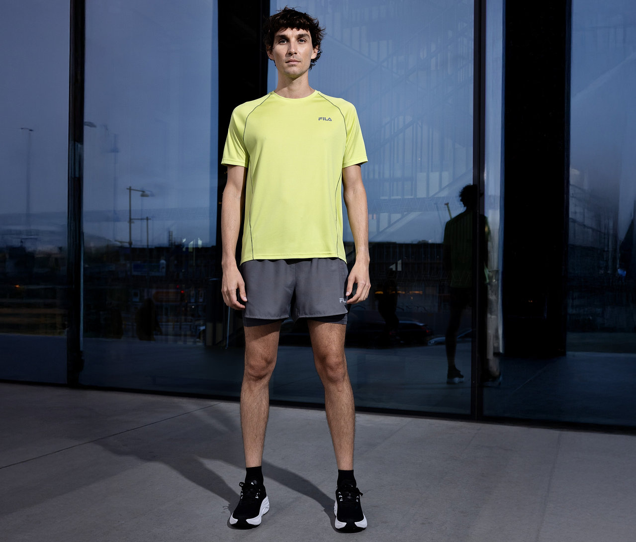 Muž stojí pred budovou a má na sebe žlté tričko Fila, sivé šortky a čierne topánky.