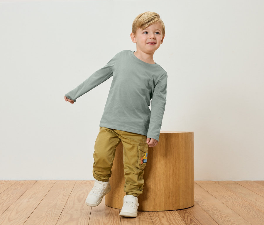 Blond chlapec v zelenom tričku s dlhým rukávom a khaki nohaviciach stojí vedľa drevenej stoličky.