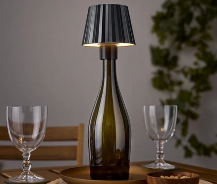 Stolná lampa v tvare vínovej fľaše stojí na drevenom stole vedľa dvoch pohárov na víno a misky s orechmi.