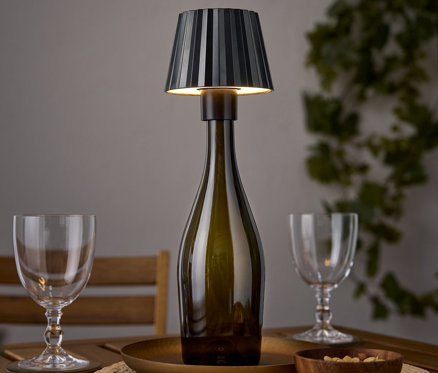 Stolná lampa v tvare vínovej fľaše stojí na drevenom stole vedľa dvoch pohárov na víno a misky s orechmi.