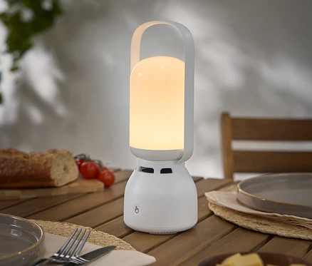 Zapnutá, prenosná, biela lampa s rúčkou stojí na drevenom stole vedľa taniera, príboru, chleba, paradajok a syra.