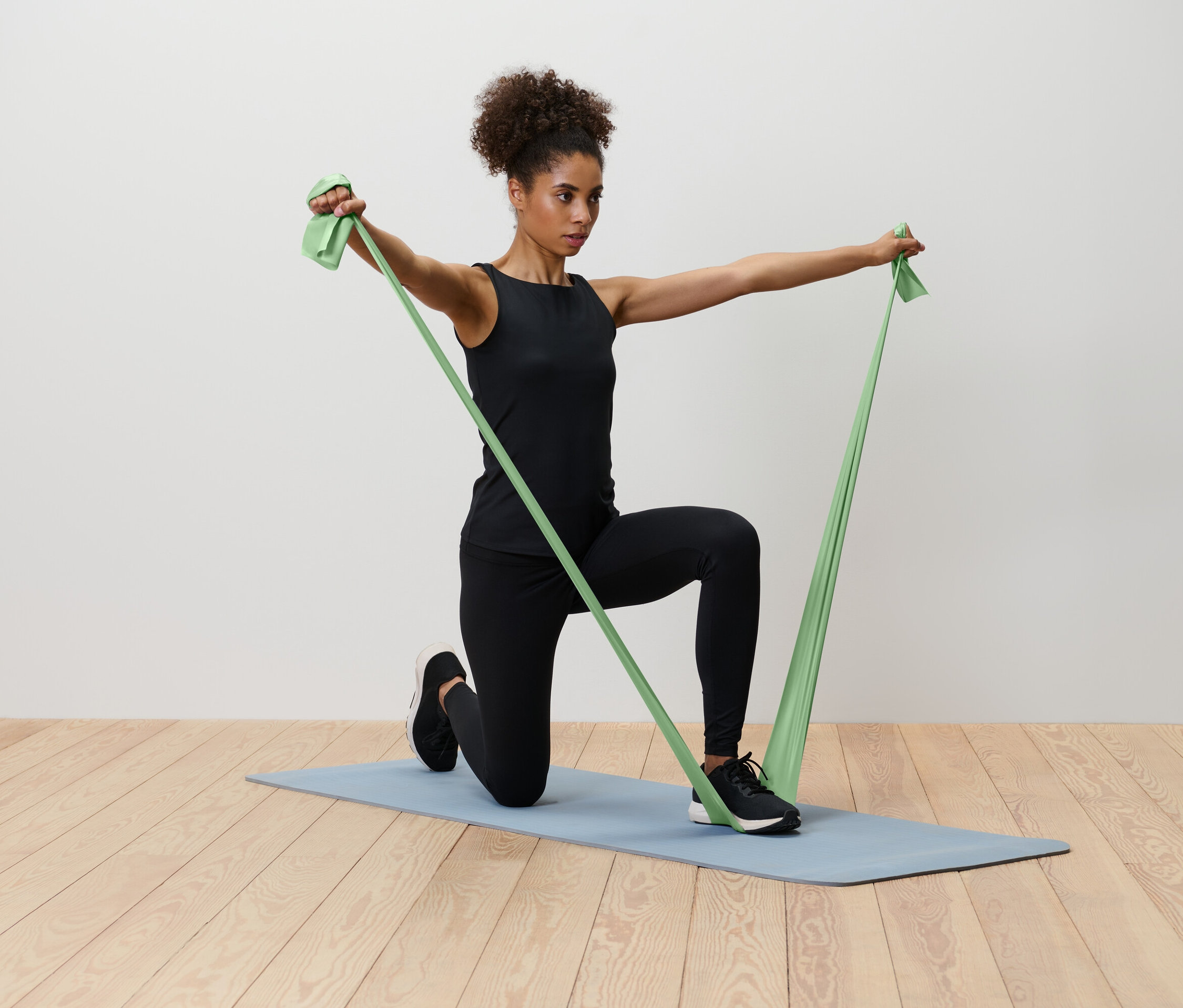 Žena kľačí na modrej podložke na jogu a naťahuje zelenú odporovú gumu.