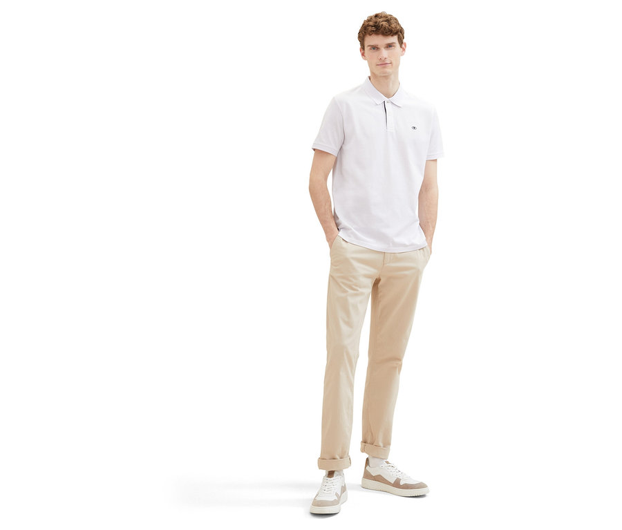 Muž v béžových nohaviciach a bielom piké polokošeli Tom Tailor. Stojí s rukami vo vreckách na bielom pozadí.