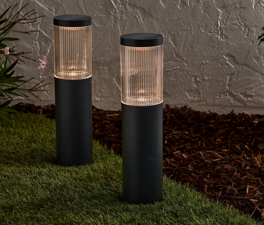 Dve čierne solárne lampy stoja na trávniku pred stenou.