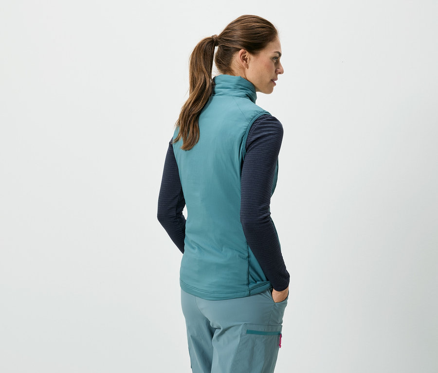 Pohľad zozadu na ženu s konským chvostom v tyrkysovej veste bez rukávov, modrej košeli s dlhým rukávom a tyrkysových nohaviciach, s rukami vo vreckách.