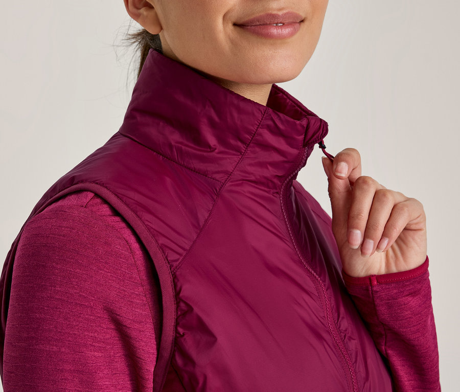 Žena v tmavoružovom tričku s dlhým rukávom a zladenej bezrukávovej prešívanej veste, zapína si zips na veste.