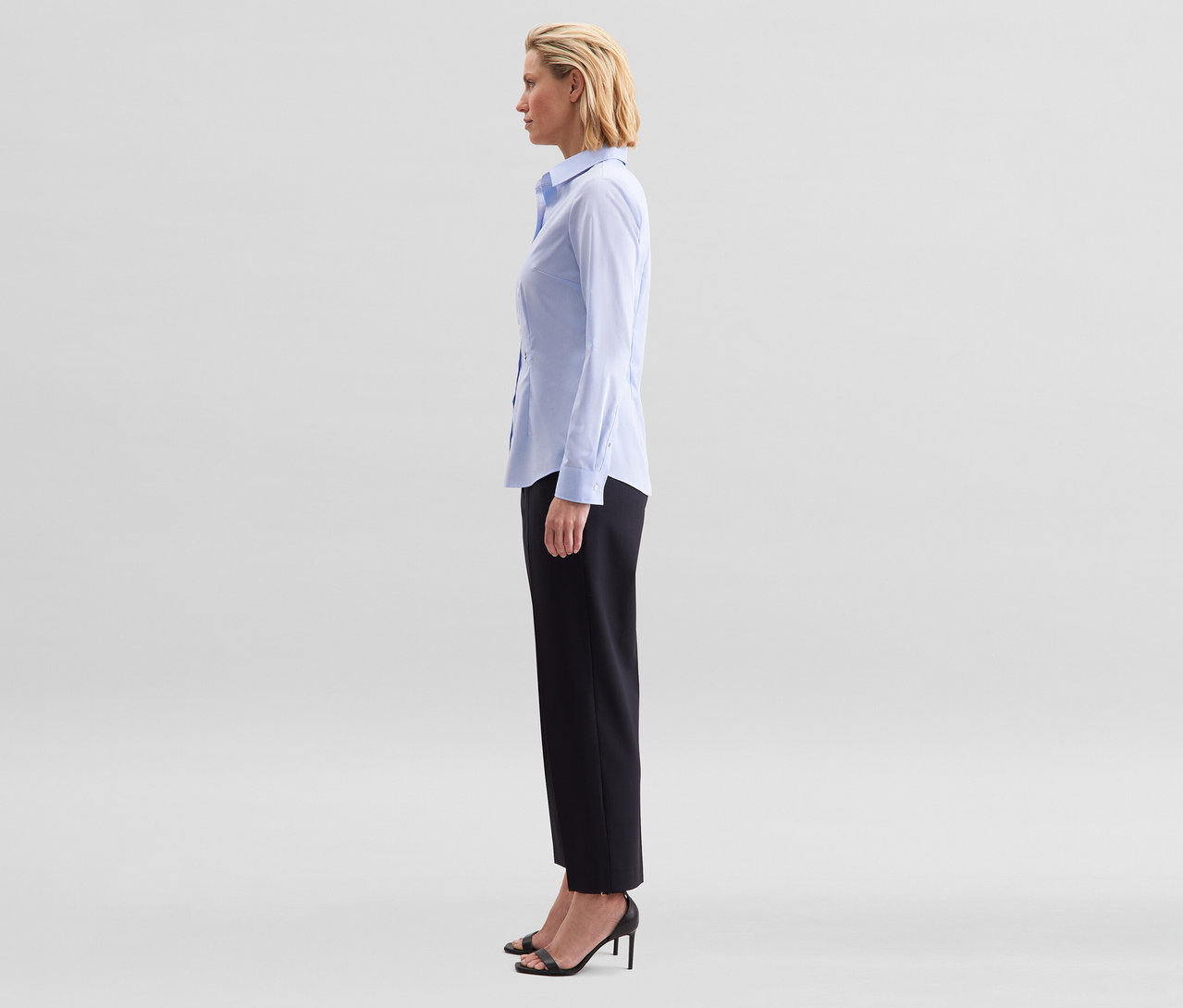 Žena stojí z profilu, má na sebe svetlomodrú blúzku Seidensticker Slim Fit non iron, čierne nohavice a čierne sandále na opätku.