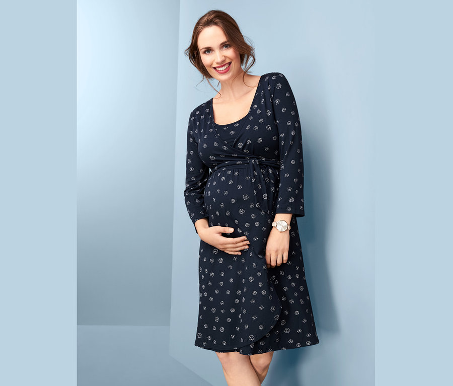 Usmievajúca sa tehotná žena drží brucho a má na sebe tmavomodré tehotenské šaty v zavinovacom vzhľade s potlačou.