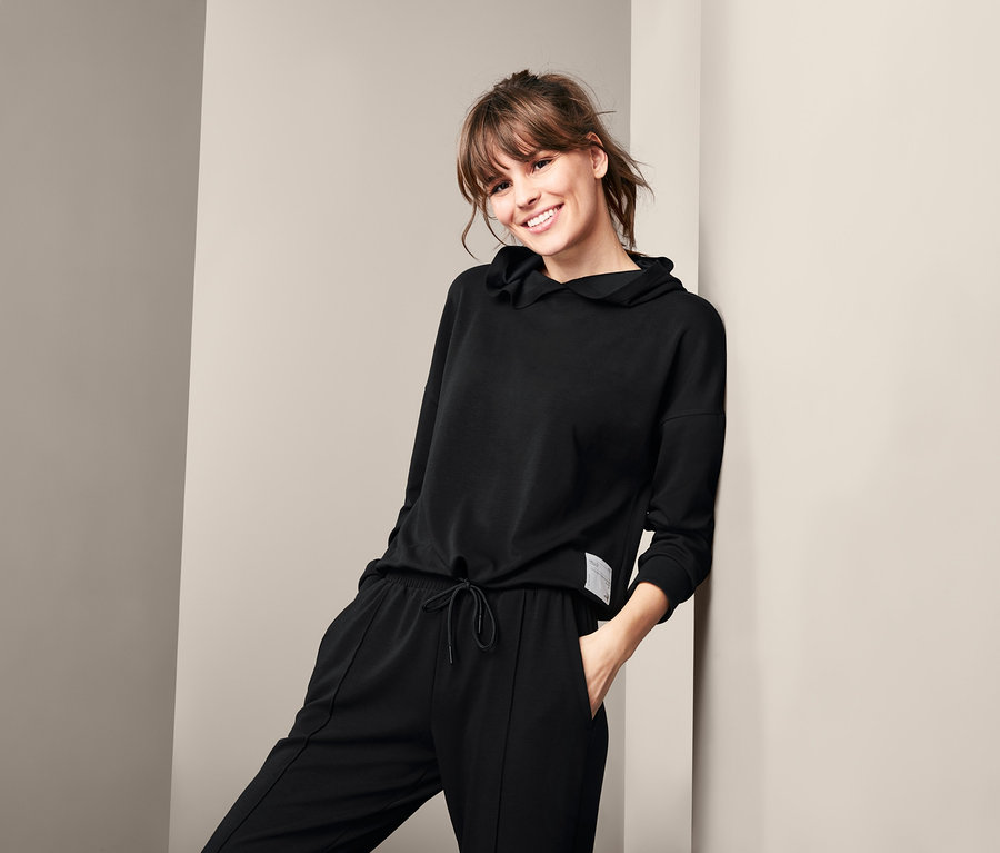 Žena sa usmieva a opiera sa o stenu v čiernej domácej mikine a nohaviciach.