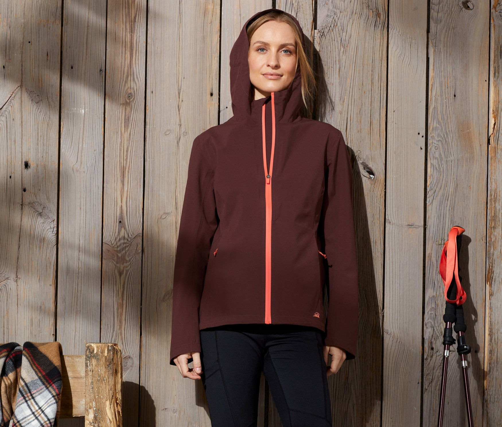 Žena stojí opretá o drevenú stenu a má na sebe funkčnú turistickú bundu s kapucňou a oranžovým zipsom.