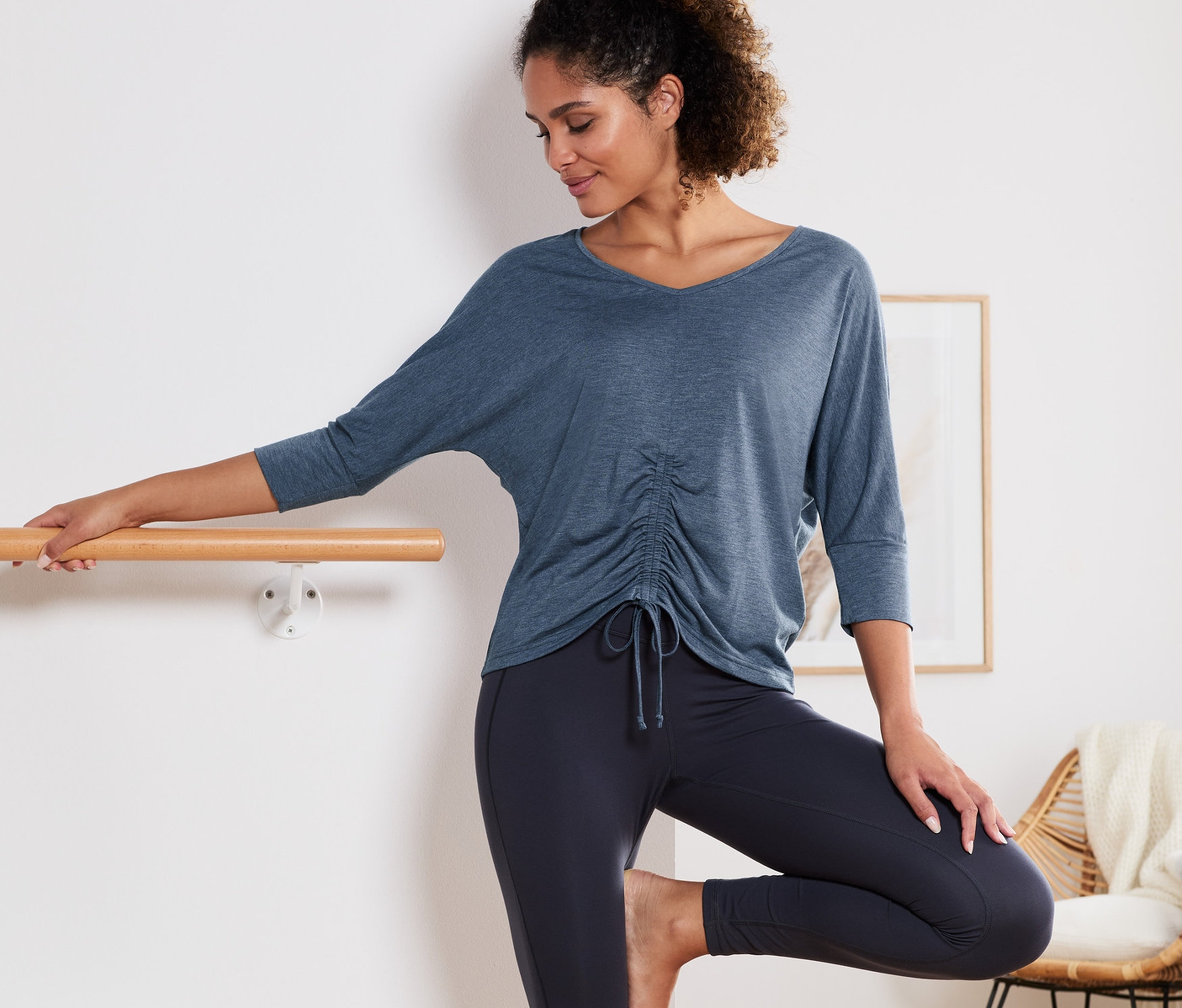 Žena pózuje pri baletnej tyči v modrom športovom tričku na jogu s trojštvrťovými rukávmi.