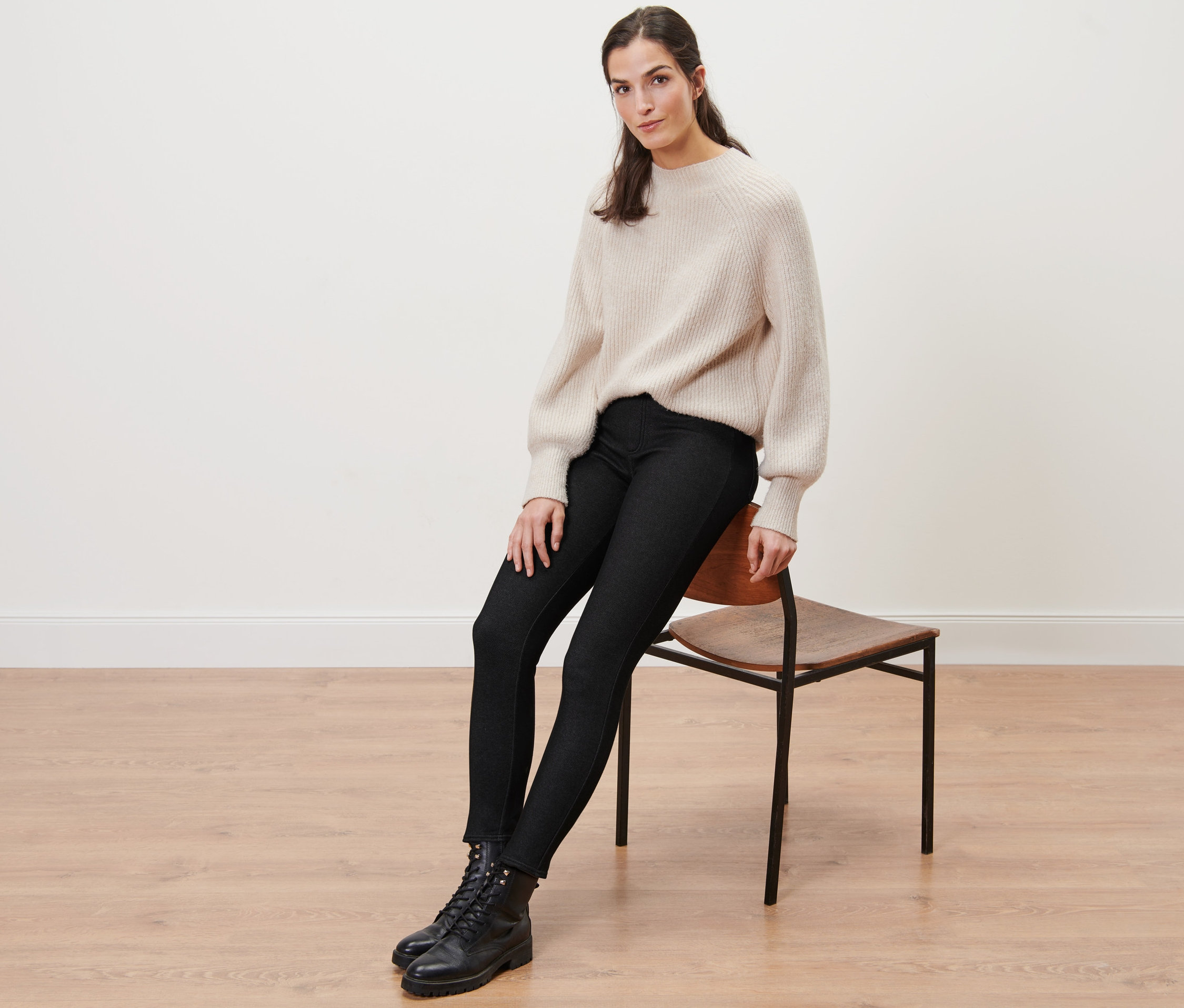 Žena sedí na stoličke a má na sebe podšité legínsy s džínsovým vzhľadom, sveter a topánky.