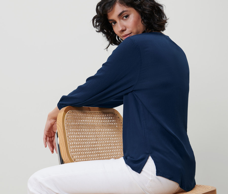 Žena sedí bokom na stoličke a má na sebe modrú tunikovú blúzku.