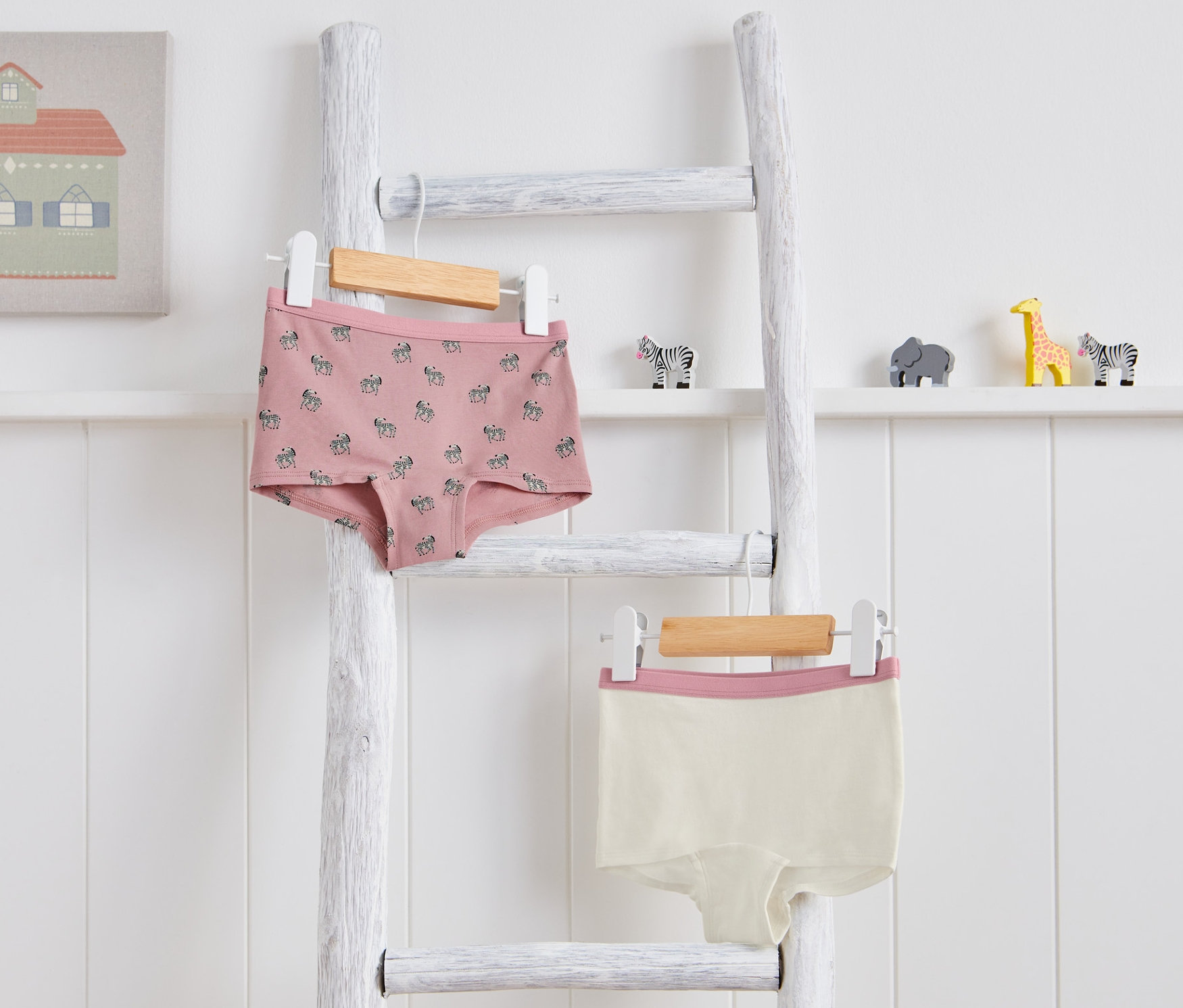 Dvoje detské nohavičky visia na bielom rebríku. Jedny sú ružové so vzorom zebry, druhé krémové s ružovým lemom.
