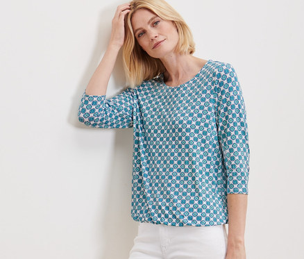 Žena pózuje opretá o stenu v blúzkovom tričku s celoplošnou potlačou a bielych nohaviciach.