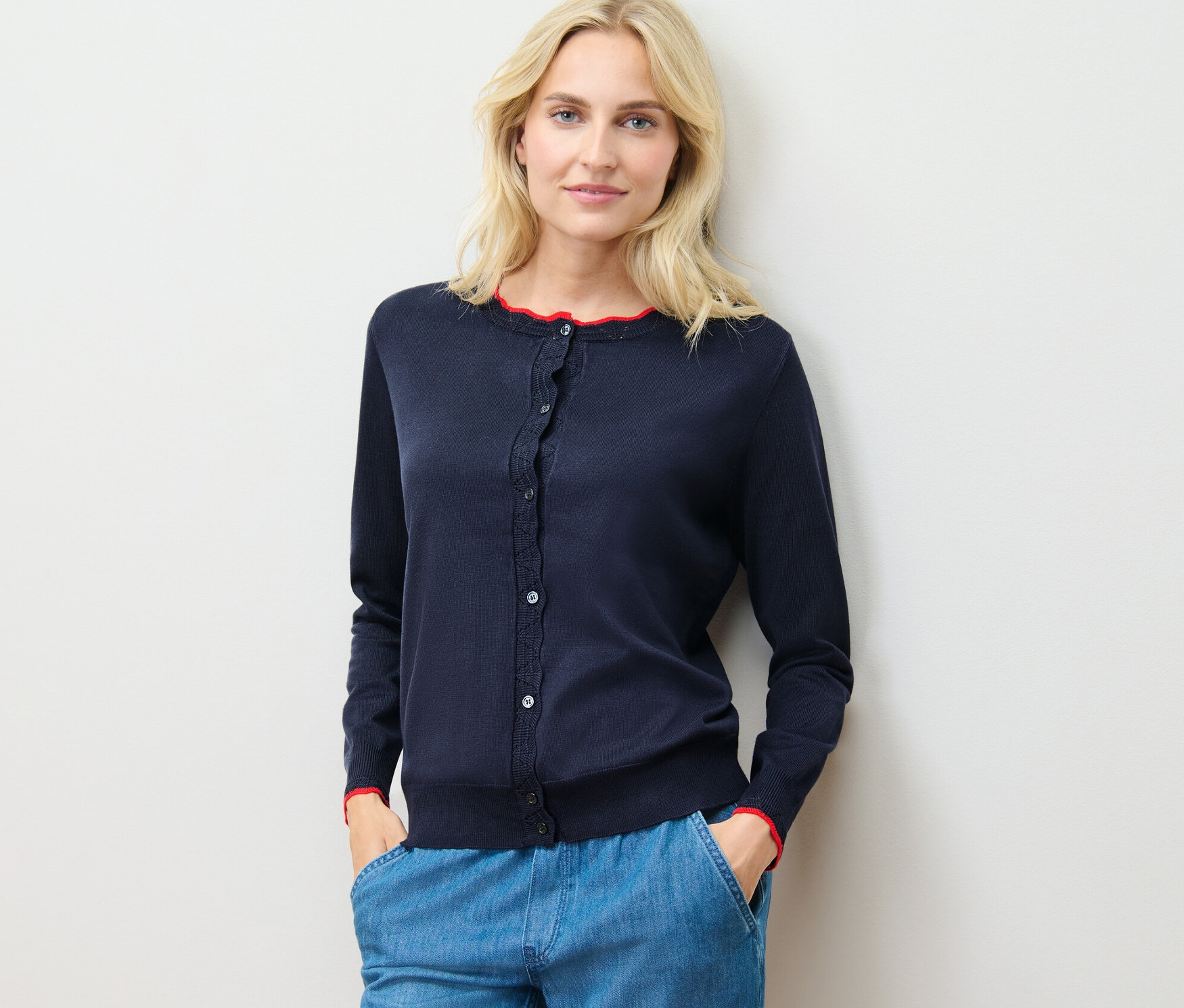 Žena má na sebe kardigán z jemného úpletu so zvlneným okrajom a modré nohavice a stojí pri stene.