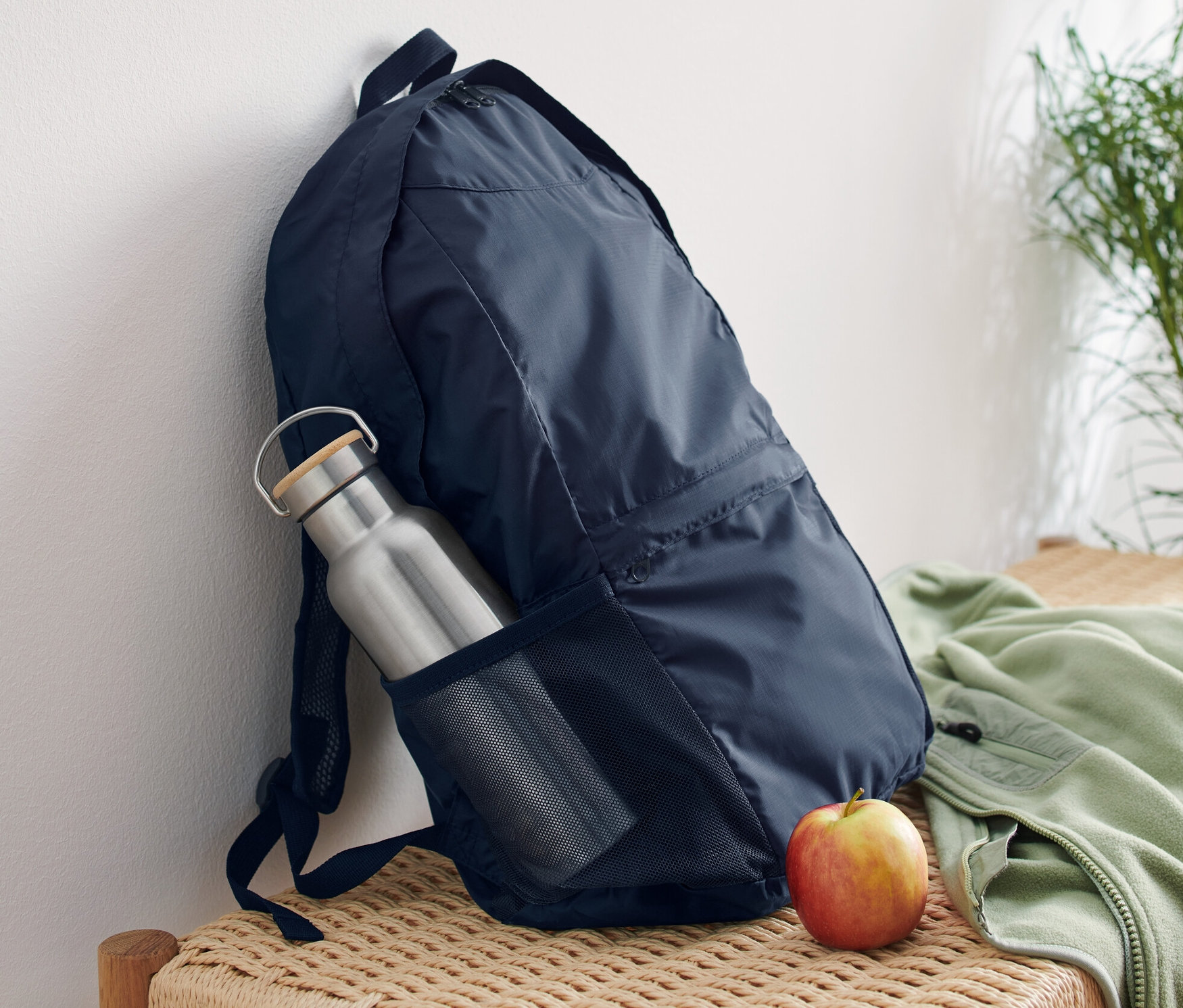 Tmavo modrý skladací ruksak opretý o bielu stenu na prútenej lavici. V bočnom vrecku ruksaku je strieborná fľaša na vodu a vedľa ruksaku leží jablko.