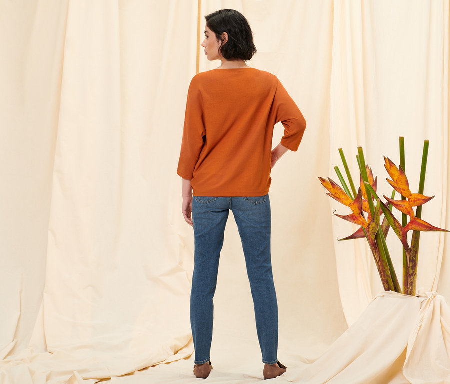 Žena stojí chrbtom k fotoaparátu a má na sebe slim džínsy Fit Emma, stredne modrá džínsovina, oranžový pulóver z jemnej pleteniny a mokasíny z velúrovej kože.