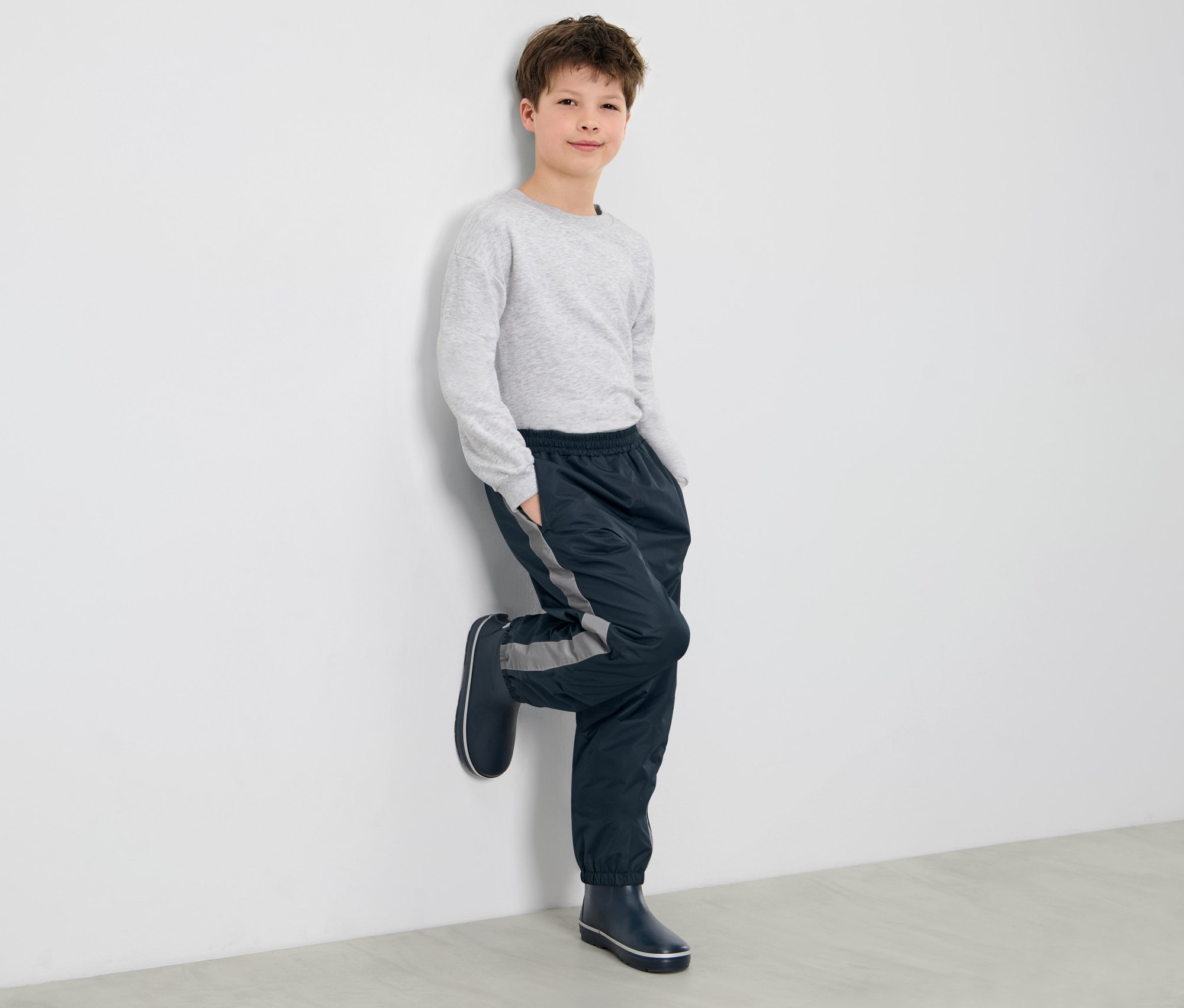 Chlapec opretý o stenu má na sebe detské nohavice do dažďa a gumáky.