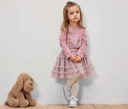 Dievčatko v ružovom tričku s dlhými rukávmi s motívom princeznej, ružovej sukni a bielych teniskách stojí pri stene. Vedľa nej sedí plyšový pes.